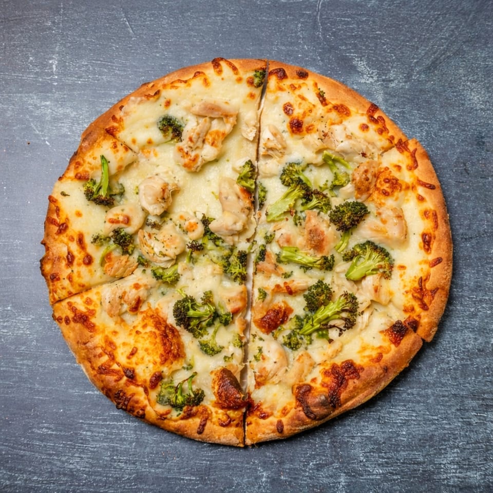 Overhead view of a white sauce pizza topped with chicken and broccoli, sliced on a dark surface.