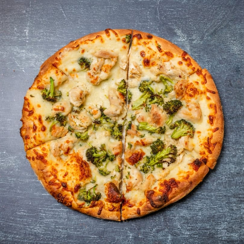 Overhead view of a white sauce pizza topped with chicken and broccoli, sliced on a dark surface.